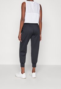 Woman wearing black jogger pants, white sleeveless top, and white sneakers standing against a plain light background, back view.