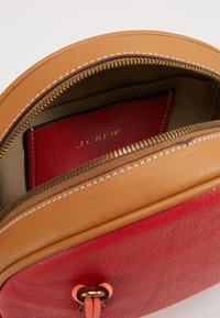 Torba na ramię z jasnobrązowej skóry z czerwonym wnętrzem, wyposażona w zamek błyskawiczny, złote okucia oraz wewnętrzną kieszeń na karty oznaczoną jako "J.Crew".