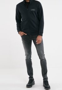 Chaqueta de cremallera azul marino con cuello alto y bolsillo frontal, combinada con jeans grises y zapatillas negras. Textura suave y diseño ajustado.