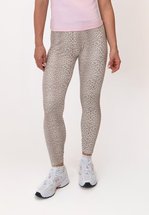 Vrouw draagt beige legging met luipaardprint, witte sportschoenen met roze accenten en een lichtroze top, staand op een effen achtergrond.
