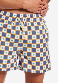 Pantaloni da nuoto con un motivo geometrico di quadrati blu, diamanti bianchi e cerchi turchesi. Caratterizzati da un girovita con cordoncino e un tessuto morbido.