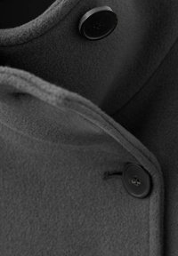 Primo piano di un soprabito in lana grigia, con una texture morbida, colletto strutturato e bottoni neri dalla finitura liscia. Dettaglio di cuciture evidenti.