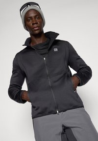 Zwarte zip-up jack met een hoge kraag, voorzakken en contrasterende naden. Gecombineerd met een zwart shirt en grijze broek.