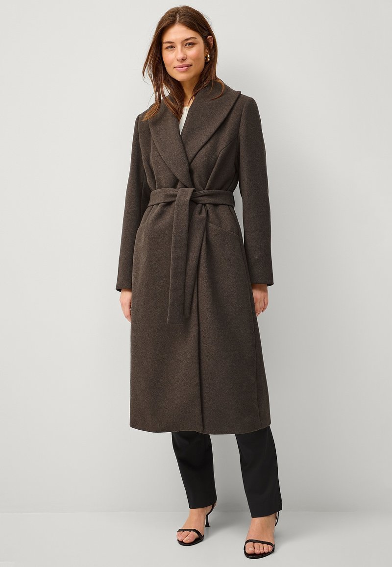 Lungo cappotto marrone scuro realizzato in tessuto testurizzato, con un ampio colletto, vita con cintura e due tasche anteriori. Indossato sopra pantaloni neri e sandali.