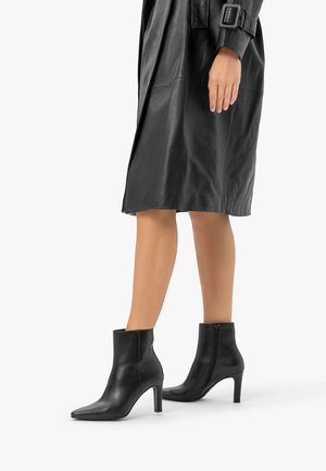 Jambes portant des bottines noires en cuir à talons et un manteau en cuir noir avec une boucle de ceinture sur la manche, sur fond blanc.