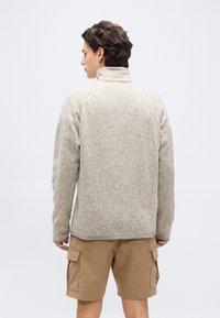 Άνδρας που κοιτάζει προς τα πίσω, με μπεζ φλις μπουφάν με ψηλό γιακά και ανοιχτό καφέ cargo σορτς, μπροστά σε απλό φόντο.