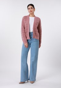 Ανοιχτό ζακέτα με μπροστινό άνοιγμα σε απαλό ροζ πλεκτό με υφασμένους ραβδώσεις, συνδυασμένο με ψηλόμεσα, φαρδιά τζιν παντελόνια σε ανοιχτό μπλε και μπεζ μυτερές γόβες.