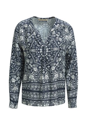 Frau steht mit einer Hand in der Tasche, trägt ein langärmliges Hemd mit schwarz-weißem Paisley-Muster und weite Hosen sowie braune Sandalen.