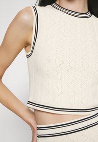 Haut tricoté crème sans manches avec bordure rayée noire porté par une personne, montrant un motif zigzag texturé et une découpe au niveau du ventre.