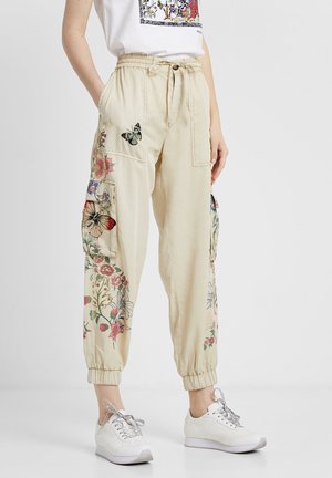 Pantalon cargo beige avec poignets et taille élastiques, présentant des imprimés floraux et papillons, porté avec des baskets blanches et un t-shirt graphique blanc.
