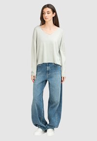 Helles grau langärmliges V-Ausschnitt-Oberteil, kombiniert mit übergroßen blauen Jeans und weißen Turnschuhen. Entspannt geschnitten mit weicher Textur.