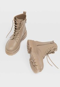 Botas de combate beige con parte superior sintética suave, cordones en la parte delantera, cremallera lateral y suelas de goma gruesa con surcos profundos para tracción.