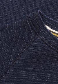 Close-up van navyblauwe gebreide stof met witte stippen, met zicht op de ronde halskraag en de stiksels van de schoudernaad.