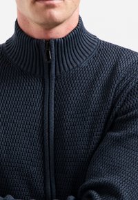 Marineblauwe zip-up sweater met een geribbeld hoge nek en een getextureerd wafelpatroon, voorzien van een rits aan de voorkant en lange mouwen.