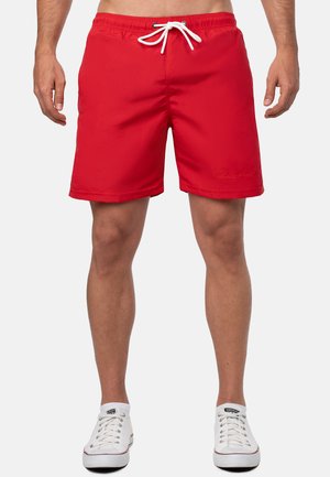 Un short de bain rouge avec une taille élastique et un cordon blanc, présentant une texture lisse et un design au-dessus du genou. Porté avec des baskets blanches.