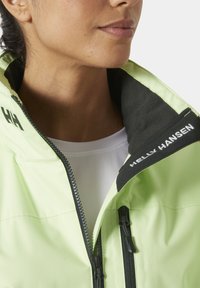 Chaqueta impermeable verde lima con ribete negro en el cuello, cremallera visible y la marca "Helly Hansen" en el cuello. Textura suave.