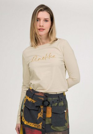 Donna con capelli biondi di media lunghezza, indossa una maglia beige a maniche lunghe e una gonna colorata con bottoni, grandi tasche e cintura.