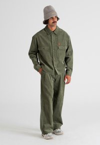 Hombre vistiendo una camisa de pana verde oliva y pantalones a juego, gorro de punto gris y zapatillas blancas, de pie con las manos en los bolsillos.