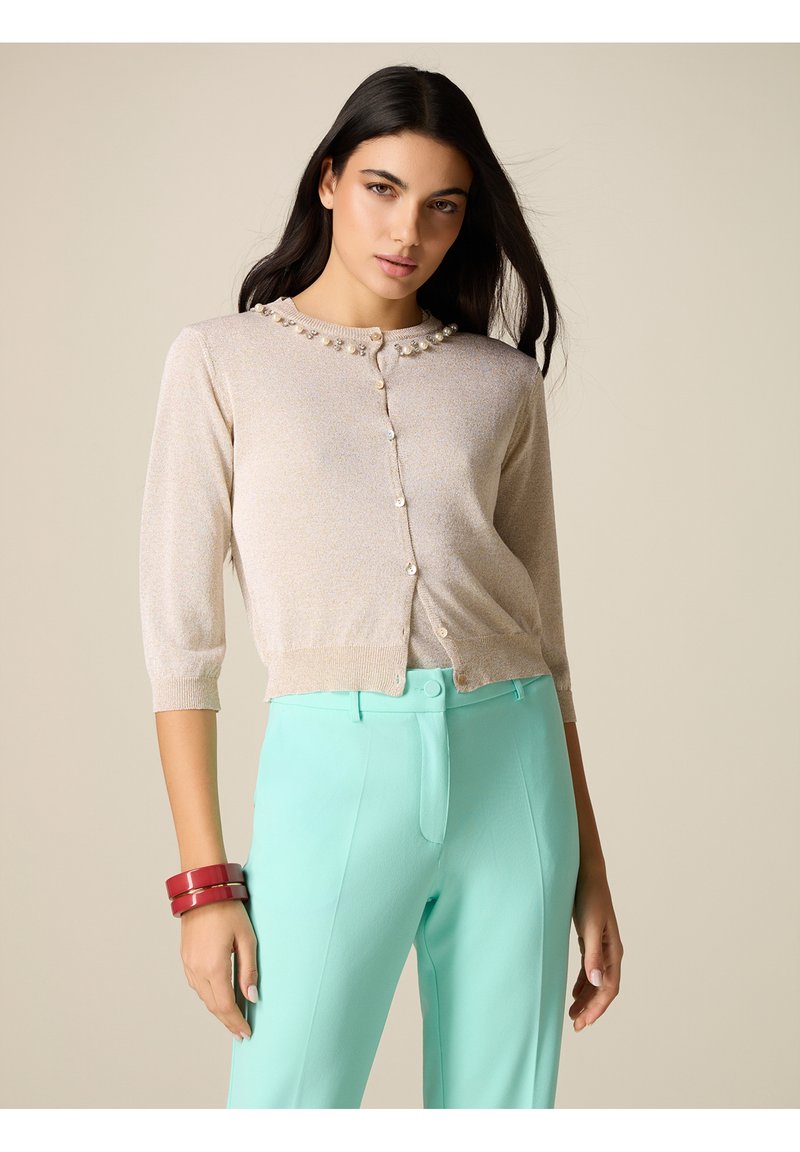 Donna con cardigan beige abbottonato con perle e pantaloni verde menta a vita alta, che indossa braccialetti rossi, posando davanti a uno sfondo beige uniforme.