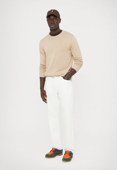 Béžový sveter, biele nohavice, farebné tenisky s oranžovými, zelenými a modrými akcentmi. Hnedá čiapka s logom.