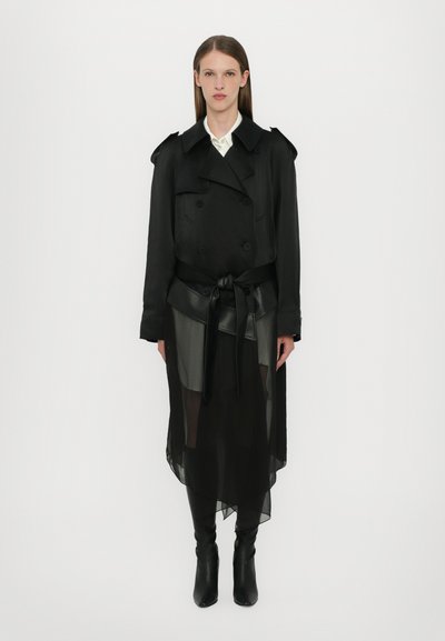Černý trench coat s dvojřadým zapínáním, se šněrovacím pasem, vrstevnatý přes průhlednou černou sukni. Obsahuje strukturovaná ramena a dlouhé rukávy.