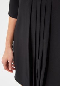 Primer plano de la mano de una persona descansando a su lado, vestida con una prenda negra con pliegues verticales en la tela.