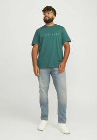 Groene katoenen t-shirt met korte mouwen, voorzien van een wit logo over de borst. Combinatie met lichtblauwe spijkerbroeken en witte sneakers.