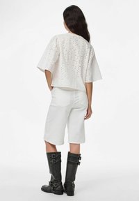 Bílý top s vyšívaným vzorem a krátkým rukávem, kombinovaný s bílými bermudami a černými vysokými botami na přezku. Jednoduchý, texturovaný design s výřezy.