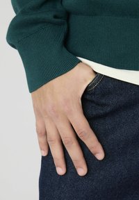 Pull en maille vert foncé avec des poignets côtelés, superposé à une chemise de couleur claire, associé à un jean en denim bleu. Main posée sur la poche.