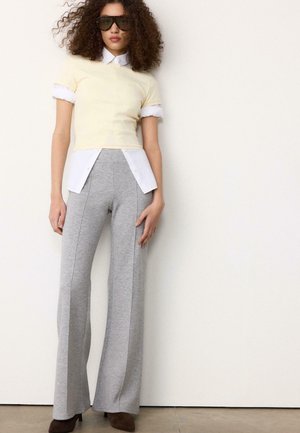 Vrouw met krullend haar, zonnebril, een gelaagde gele en witte top, grijze wijd uitlopende broek en bruine schoenen met hak, staand tegen een witte muur.