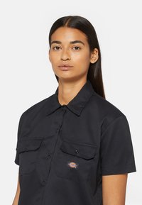 Schwarzes Kurzarmhemd mit Knopfleiste, zwei Brusttaschen und einem Logopatch. Glattes Material mit einem klassischen Kragen.
