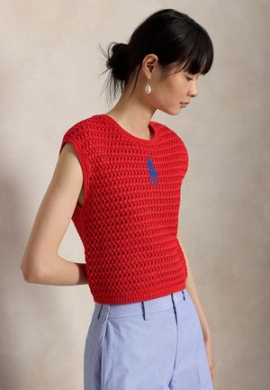 Fekete haját kontyba tűző nő, vörös ujjatlan kötött felsővel, kék logóval és világoskék, magasított derekú nadrággal, oldalirányban áll.