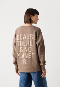 Brun oversized genser med hevet, kremfarget tekst på baksiden: "FORDI DET IKKE FINNES NOE PLANET B." Løs passform med ribbestrikkede mansjetter.