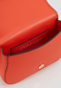 Sac en cuir orange Calvin Klein avec une texture lisse, une forme incurvée, un bord noir et une fermeture à bouton en métal doré. L'intérieur comporte un logo.
