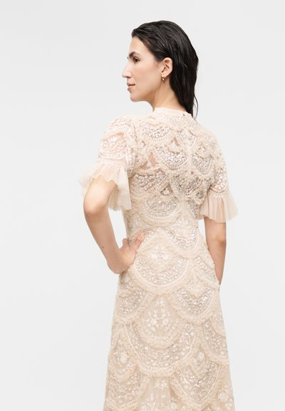 Γυναίκα με σκούρα μαλλιά με υγρή όψη φοράει εκρού δαντελένιο φόρεμα με φουσκωτά μανίκια και μοτίβα με φλοράλ παγιέτες, ποζάροντας με το χέρι στη μέση, στραμμένη προς τα αριστερά.