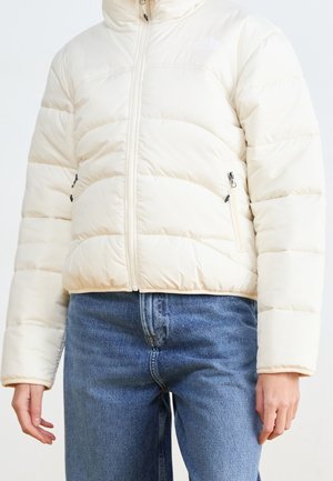 Personne portant une doudoune couleur crème avec une fermeture éclair à l'avant et un jean bleu en denim, debout avec les mains détendues le long du corps.