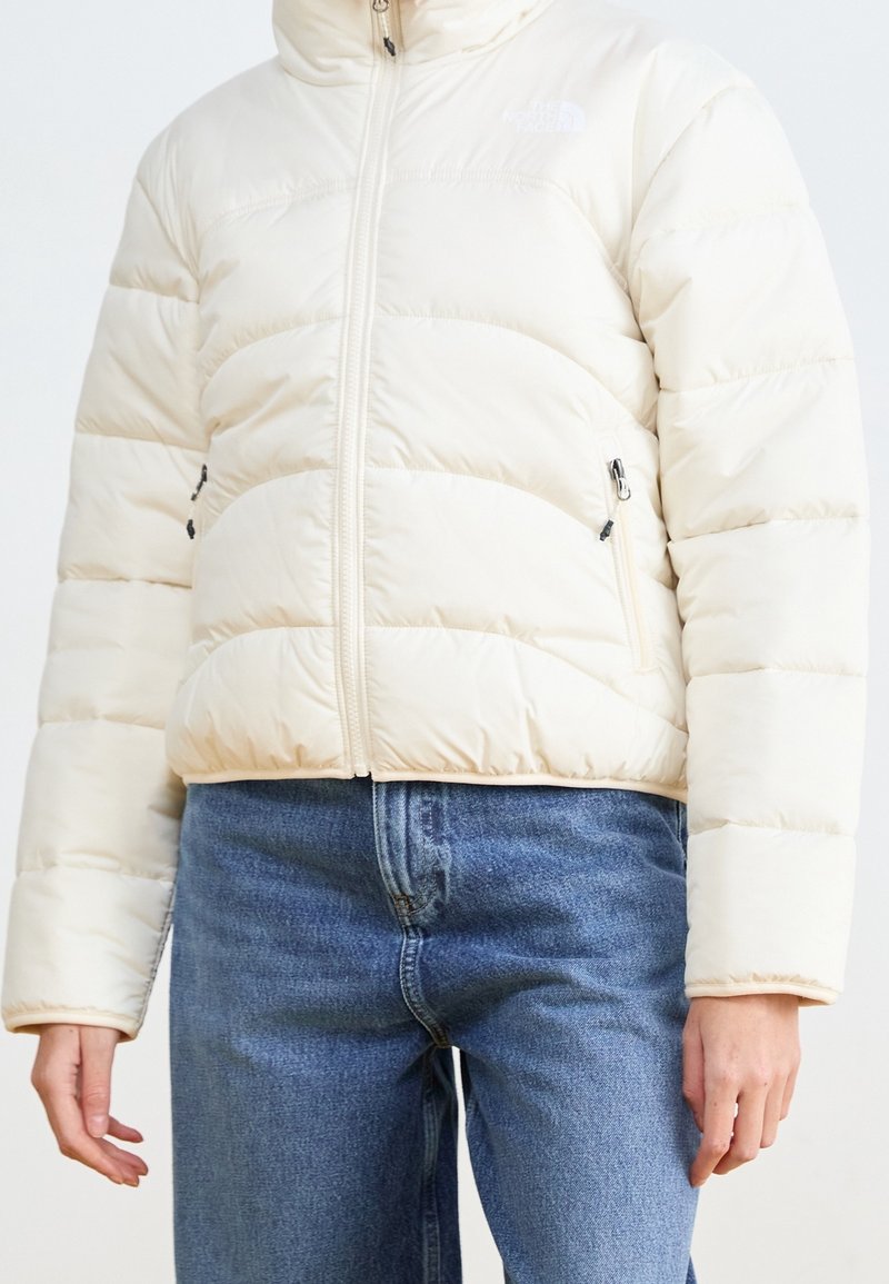 Personne portant une doudoune couleur crème avec une fermeture éclair à l'avant et un jean bleu en denim, debout avec les mains détendues le long du corps.