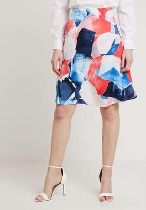 Mujer con una falda a la altura de la rodilla con estampado abstracto colorido, blusa blanca y sandalias blancas de tacón alto, de pie sobre un fondo liso.