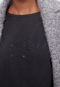 Camiseta de algodón negra con diseño de texto en relieve. Cárdigan gris suave y texturizado caído sobre el hombro, añadiendo contraste.