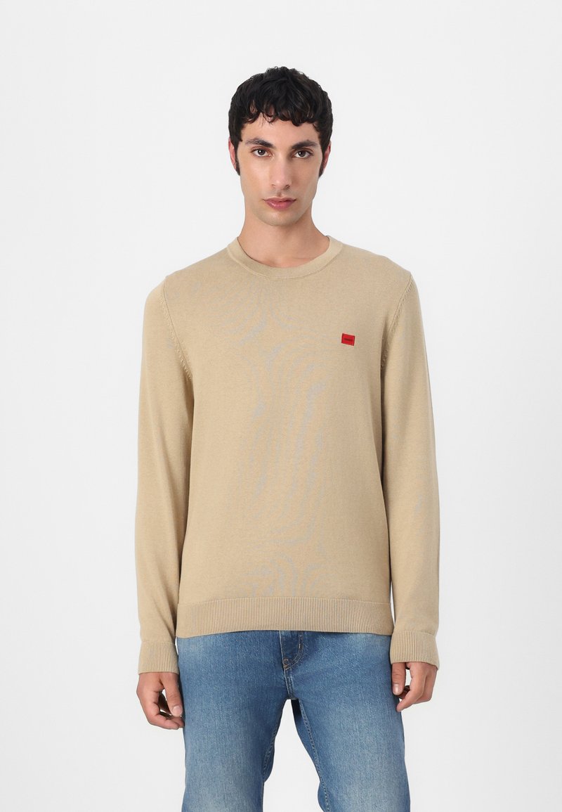 Beige Pullover mit langen Ärmeln aus einem weichen Stoff, der eine subtile Textur aufweist, gerippte Bündchen hat und ein kleines rotes rechteckiges Logo auf der Brust trägt.