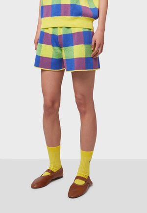 Pantaloni corti in maglia con un motivo a scacchi multicolore di blu, verde e giallo. Presentano una vita con cordoncino e un bordo giallo brillante. Abbinati a calze gialle e scarpe marroni.