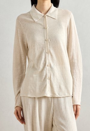 Femme portant une chemise beige à manches longues boutonnée avec un col et un pantalon crème à larges rayures fines, montrée de la poitrine à la cuisse.