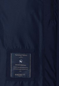 Donkerblauwe stof met een genaaide label waarop staat "Technische Stoffen door Hackett, Waterafstotend, beschermt tegen lichte regenbuien en wind."