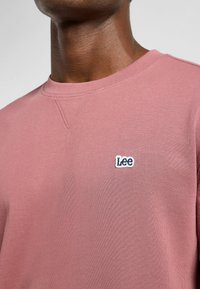 Vaaleanpunainen collegepaita, joka on valmistettu pehmeästä kankaasta. Paitassa on pyöreä pääntie, ribbireunat hihoissa ja pieni valkoinen sekä sininen brodeerattu "Lee" -logo rinnassa.