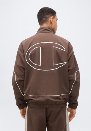 Man wearing brown tracksuit with large white outlined logo on back and white piping details on sleeves and hem.