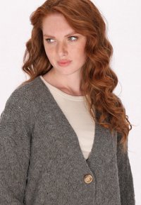 Cardigan grigio a maglia con motivo a diamante, scollatura a V e chiusura con un singolo bottone in legno. Indossato sopra un top beige chiaro.