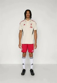 Ανδρική φανέλα της Liverpool FC με κρέμα μπλούζα και κόκκινες λεπτομέρειες, μαύρες πλαϊνές λωρίδες και κόκκινες βερμούδες. Οι κάλτσες είναι λευκές με μαύρες ρίγες.