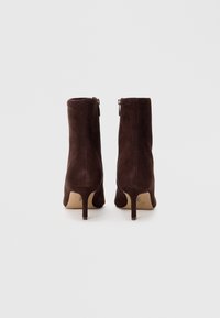 Botas de tobillo de ante marrón con un tacón delgado y un zipper lateral. Textura suave con un diseño limpio y minimalista. La altura llega a la mitad de la pantorrilla.