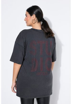 Mujer con el pelo oscuro recogido en una coleta, lleva pendientes de aro dorados y una camiseta negra de gran tamaño con "STUDIO UNTOLD" en rojo en la espalda.