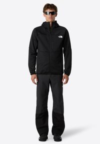 Schwarze Fleecejacke mit Reißverschluss, hohem Kragen und Logo auf der Brust; kombiniert mit schwarzen Hosen mit Color-Blocking-Design; getragen mit Sonnenbrille.